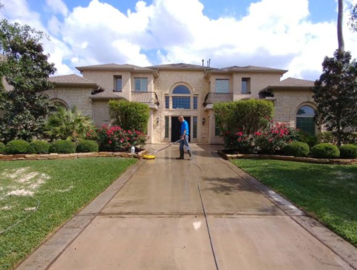 Worker surface-cleaning a long concrete driveway in front of a large stone home